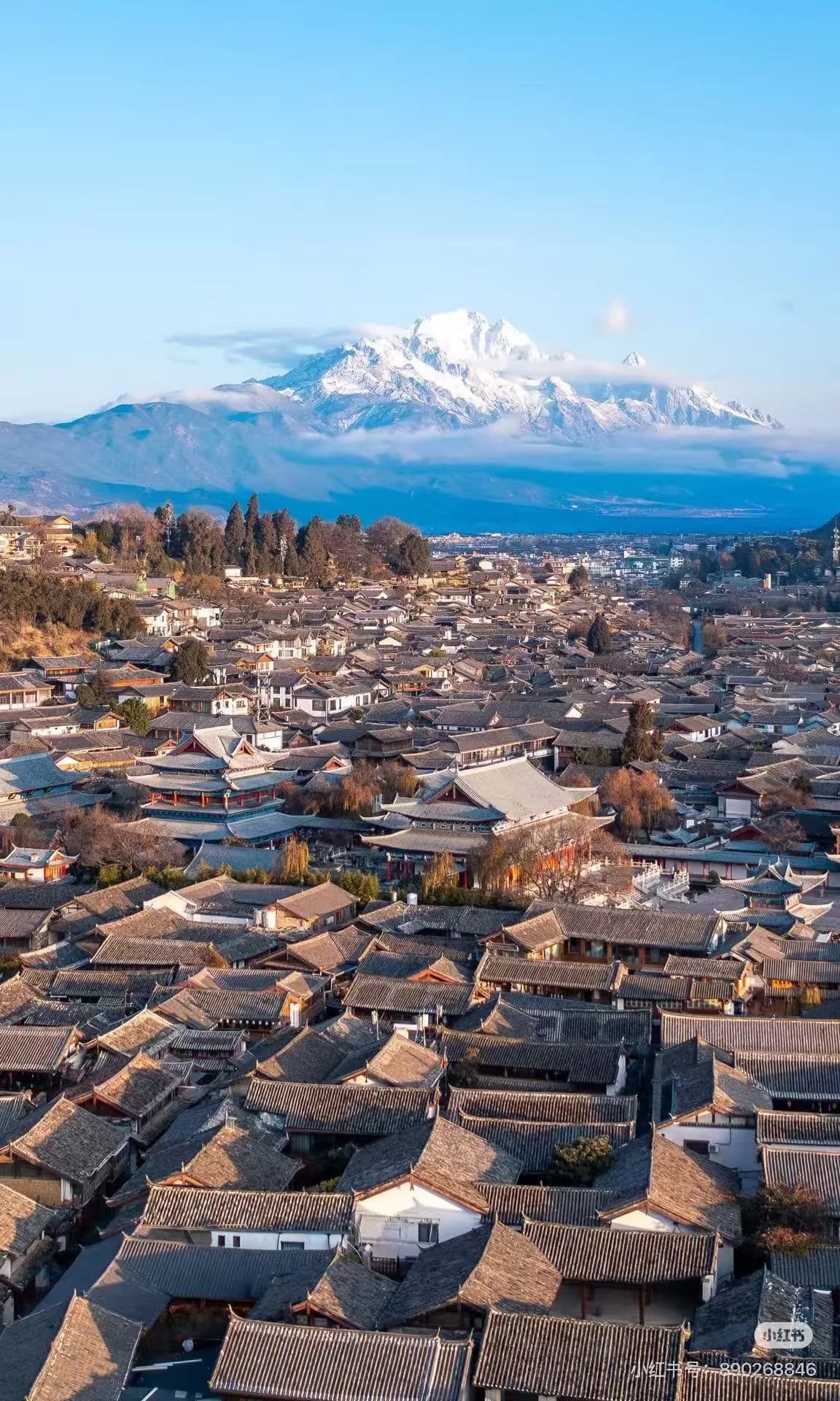 Lijiang Old Town at dusk with traditional Naxi wooden architecture, stone bridges over canals, and Jade Dragon Snow Mountain in the background