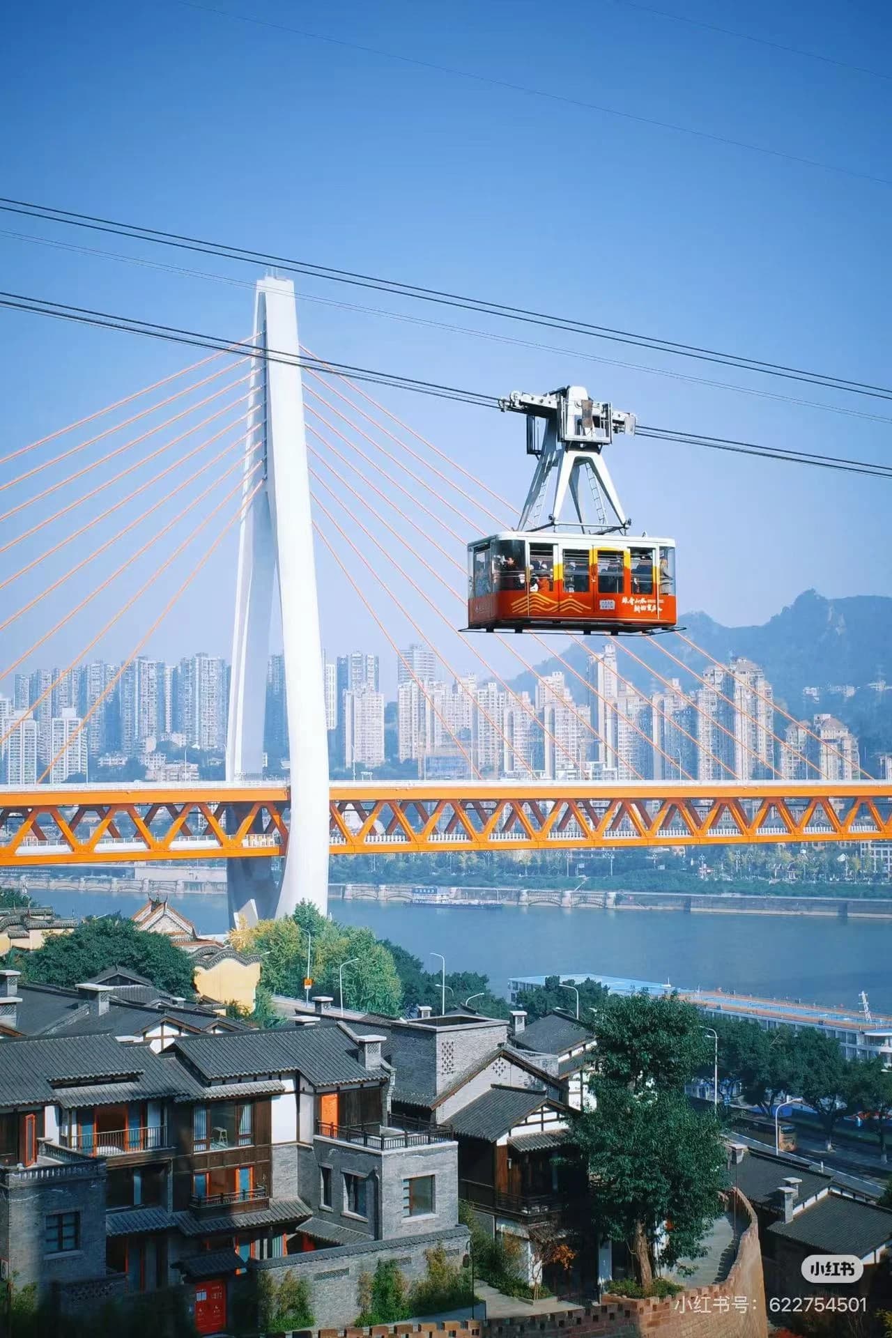 A red and orange cable car crossing the Yangtze River, with a large white suspension bridge and the city skyline in the background.